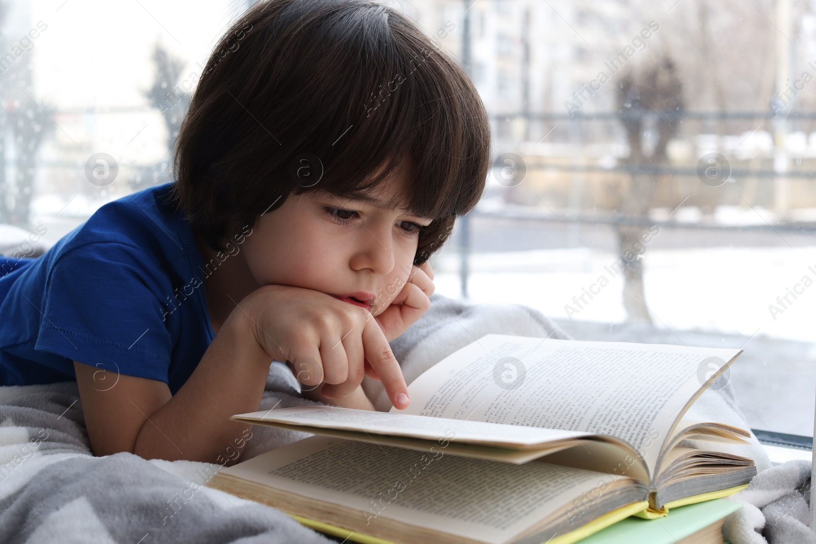 Photo of Cute little boy reading book near window at home