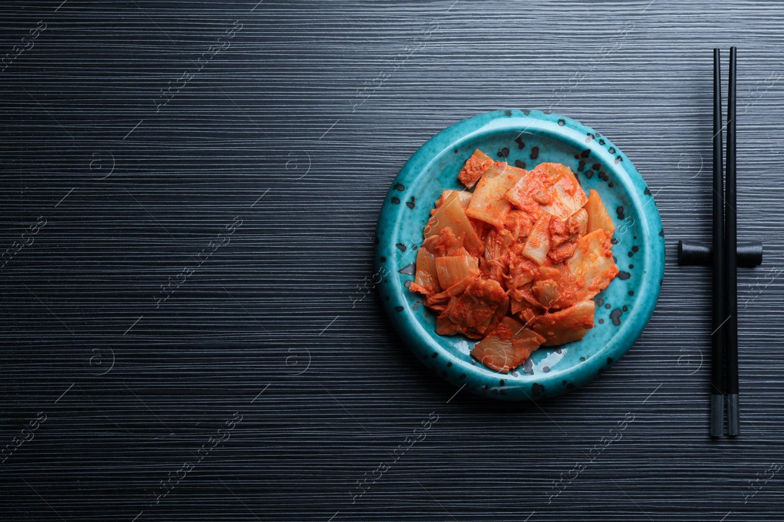 Photo of Plate of spicy cabbage kimchi and chopsticks on black wooden table, flat lay. Space for text