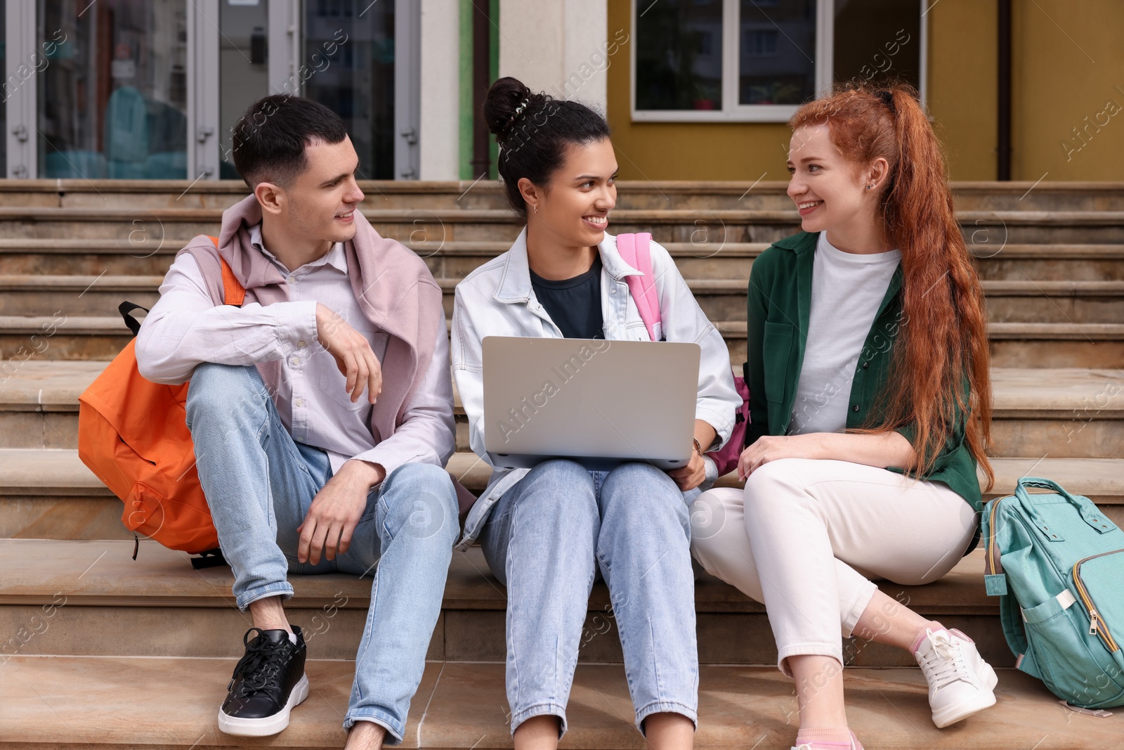 Photo of Happy young students studying together with laptop on steps outdoors