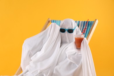 Photo of Person in ghost costume and sunglasses with glass of drink relaxing on deckchair against yellow background