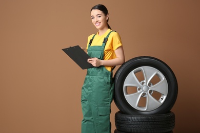 Female mechanic in uniform with car tires and clipboard on color background