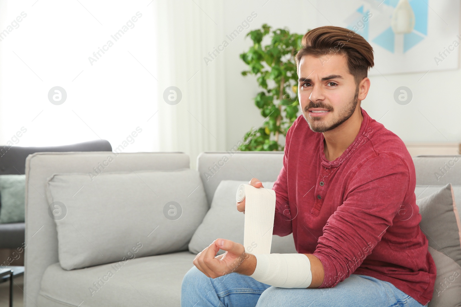 Photo of Young man applying bandage on injured arm at home, space for text. First aid