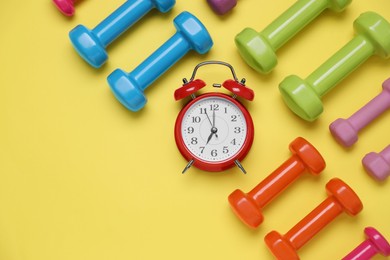 Photo of Alarm clock and dumbbells on yellow background, flat lay. Morning exercise