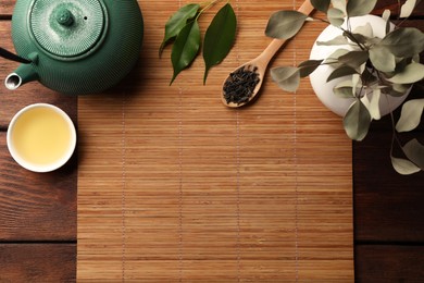 Flat lay composition with bamboo mat, dry tea leaves and teapot on wooden table, space for text