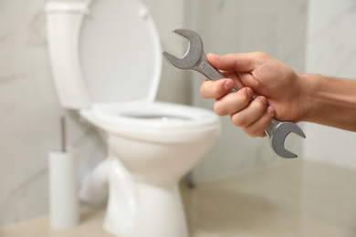 Photo of Professional plumber holding wrench near toilet bowl in bathroom, closeup