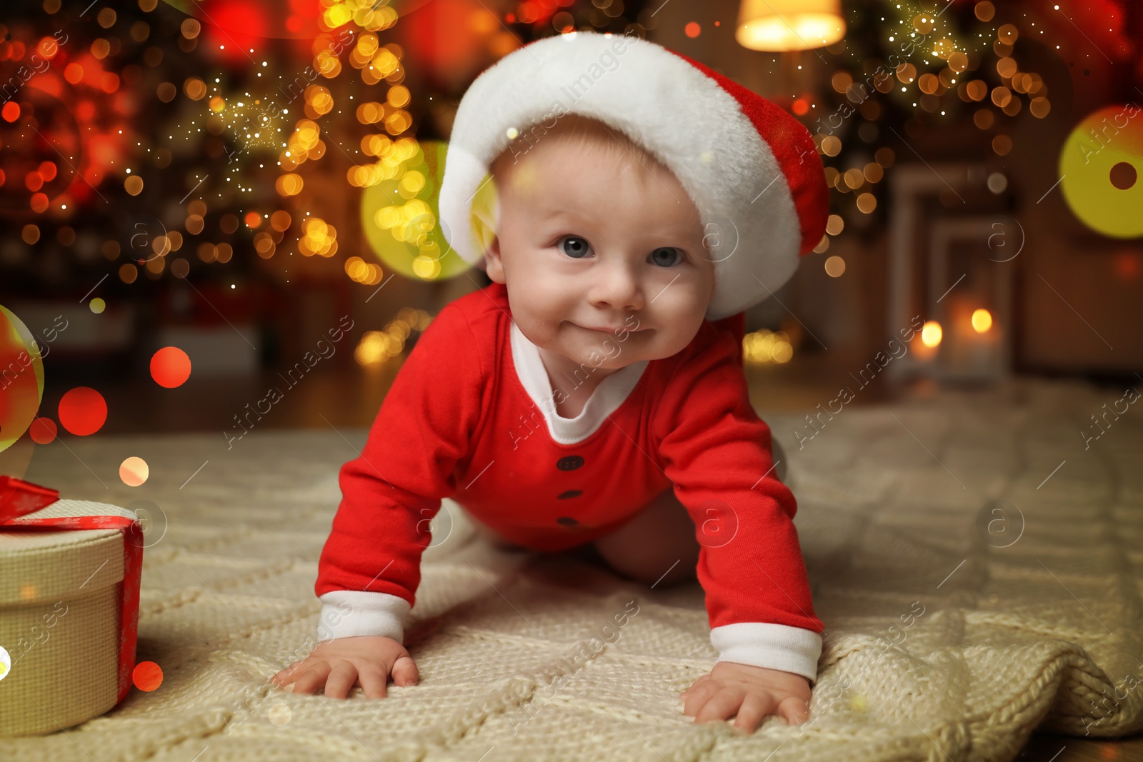 Image of Cute baby in Santa hat with Christmas gift on floor at home