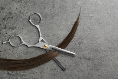 Flat lay composition with strand of brown hair and thinning scissors on grey background. Hairdresser service