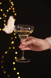 Photo of Woman holding glass of cocktail decorated with tasty cotton candy on black background with blurred lights, closeup