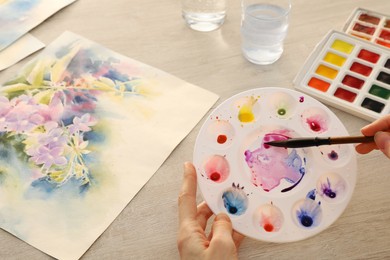 Photo of Woman painting flowers with watercolor at white wooden table, above view. Creative artwork