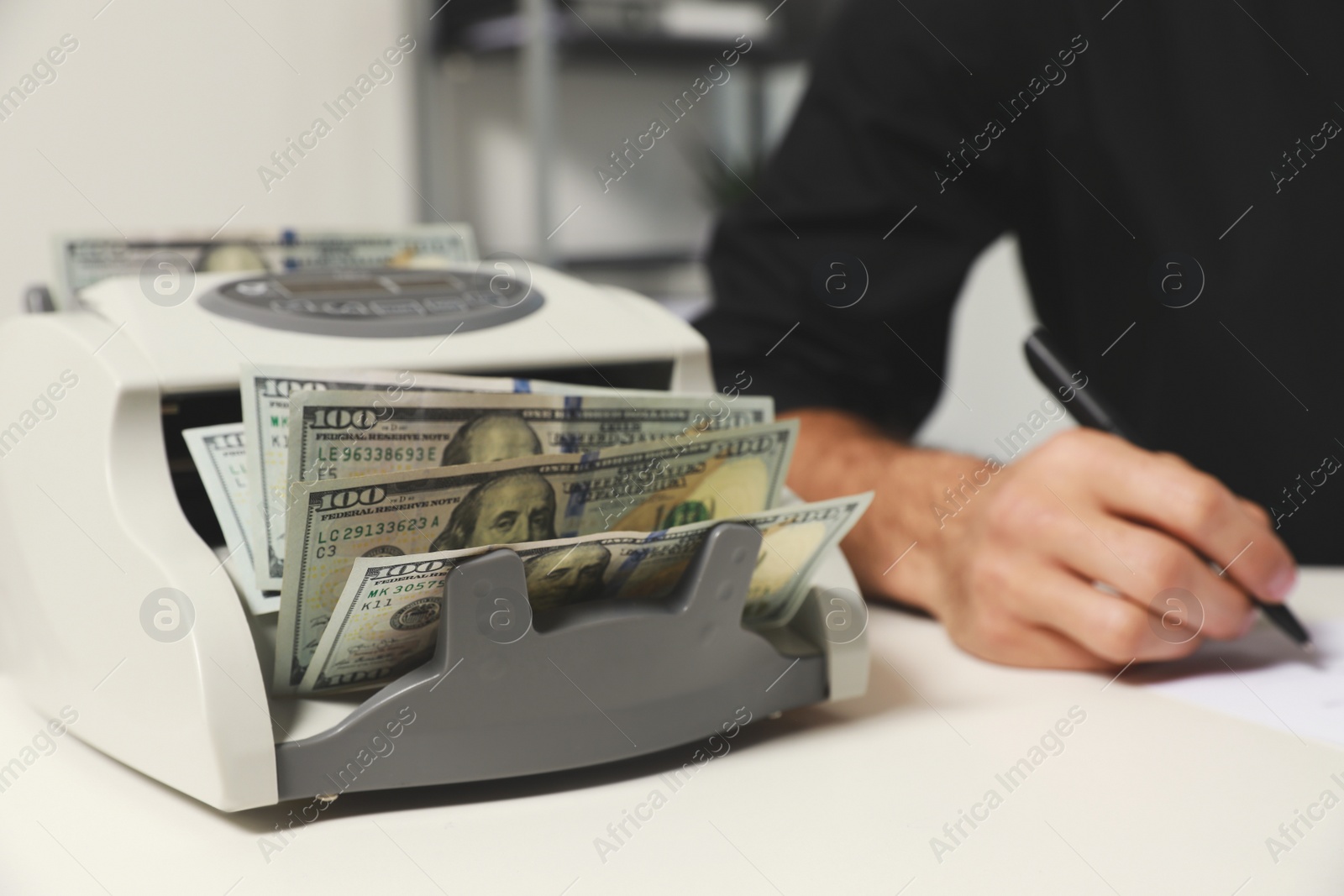 Photo of Modern banknote counter with money and blurred view of man working at white table indoors