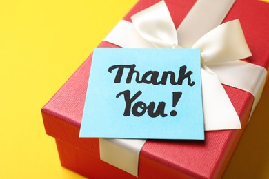 Photo of Gift box and light blue paper note with phrase Thank You on yellow background, closeup