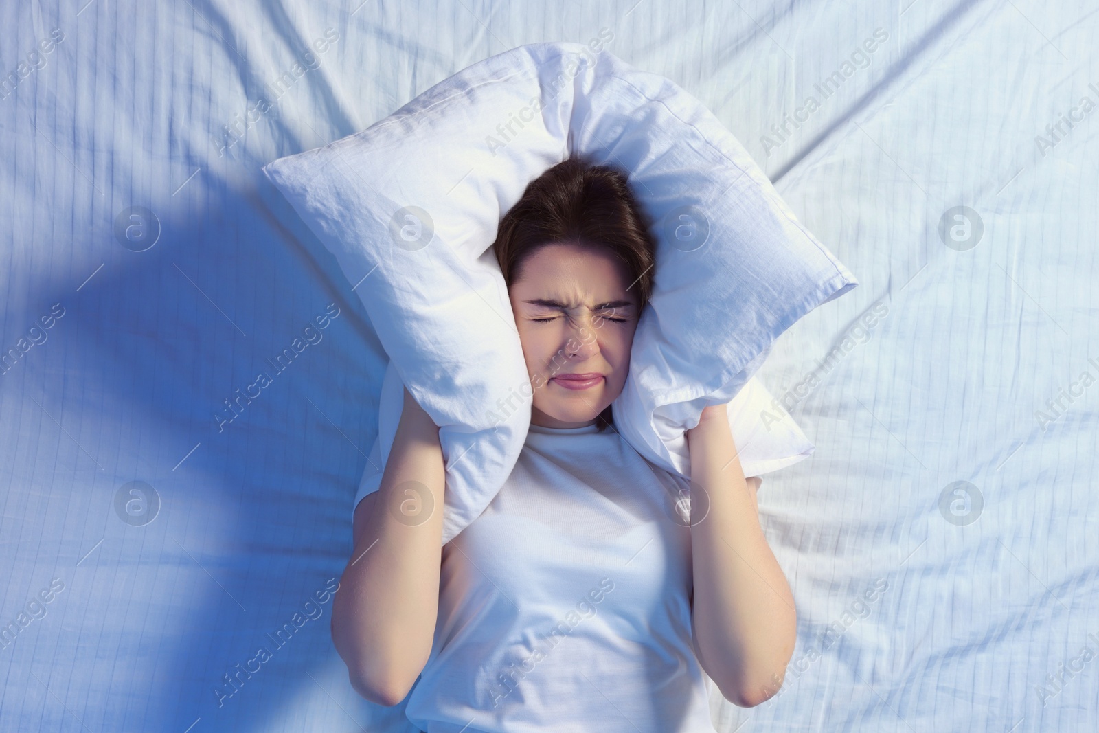 Photo of Irritated young woman covering her ears with pillow in bed, top view. Insomnia problem