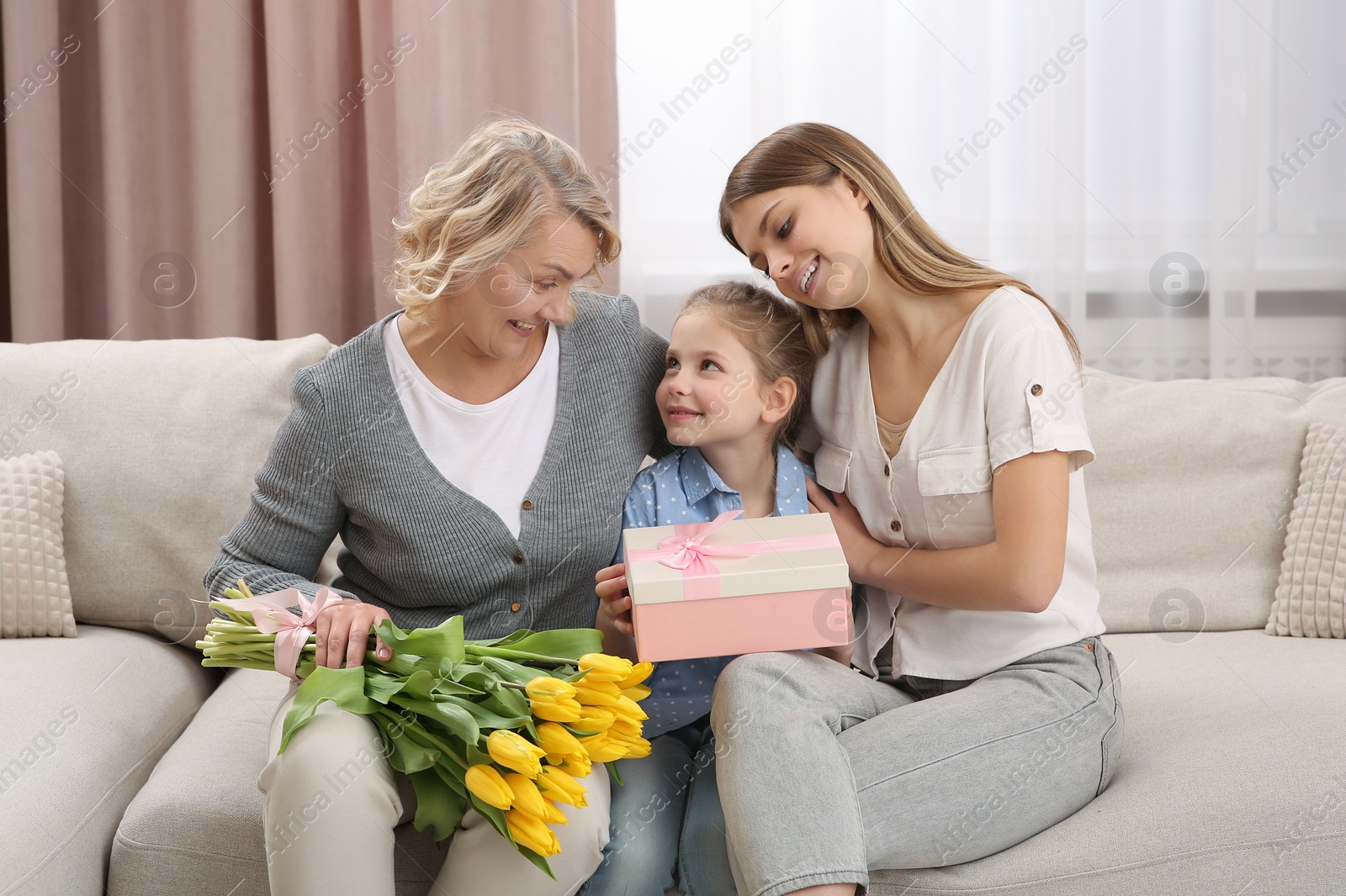 Photo of Little girl congratulating her mom and granny with flowers and gift at home. Happy Mother's Day