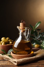 Photo of Glass jug of oil, ripe olives and green leaves on wooden table