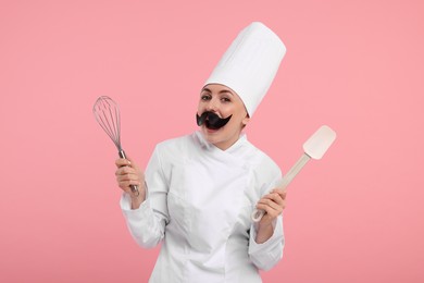 Photo of Funny professional confectioner in uniform holding whisk and spatula on pink background