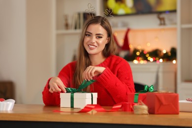 Beautiful young woman in deer headband decorating Christmas gift at table in room