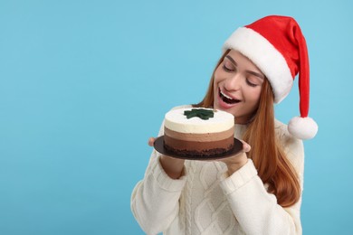 Young woman in Santa hat with tasty cake on light blue background, space for text