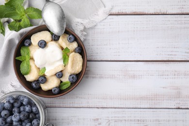 Photo of Bowl of tasty lazy dumplings with blueberries, sour cream and mint leaves on white wooden table, flat lay. Space for text