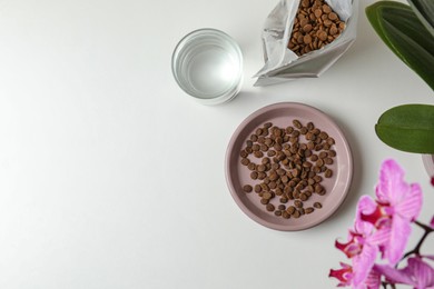 Photo of Dry cat food, glass of water and flowers on white table, flat lay. Space for text