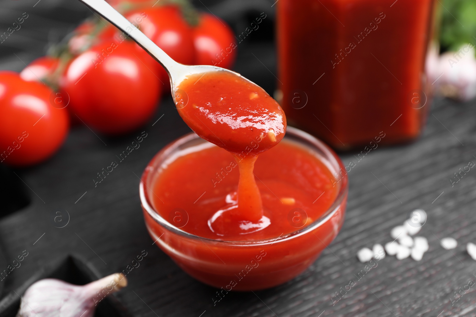 Photo of Taking tasty ketchup with spoon from bowl at black wooden table, closeup. Tomato sauce