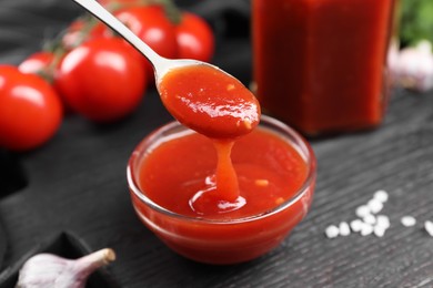 Photo of Taking tasty ketchup with spoon from bowl at black wooden table, closeup. Tomato sauce