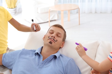 Little children painting their father's face while he sleeping on couch at home. April fool's day prank