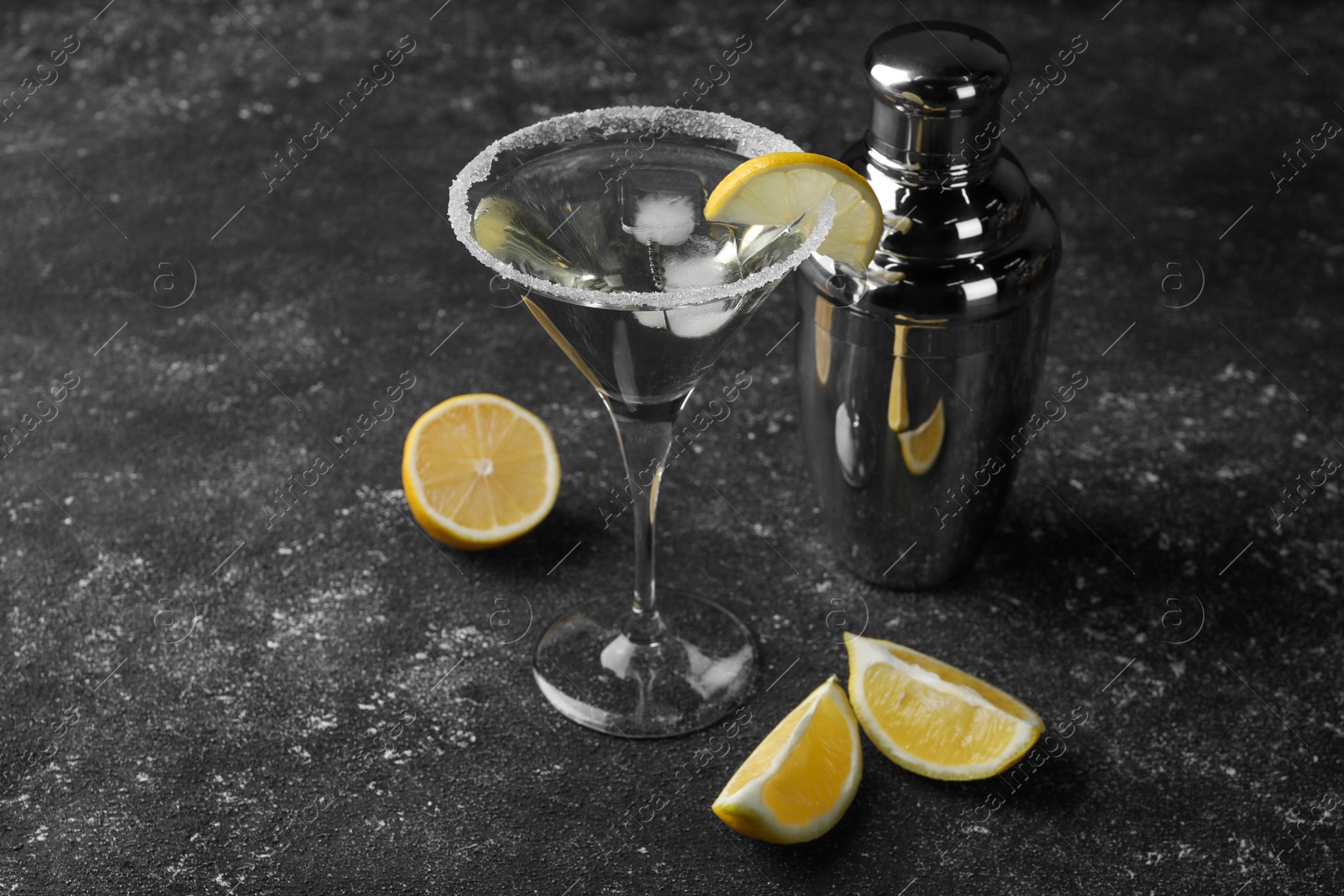 Photo of Martini glass of refreshing cocktail decorated with lemon slice and sugar near shaker on black textured table