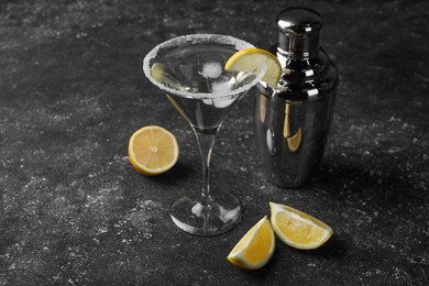 Photo of Martini glass of refreshing cocktail decorated with lemon slice and sugar near shaker on black textured table