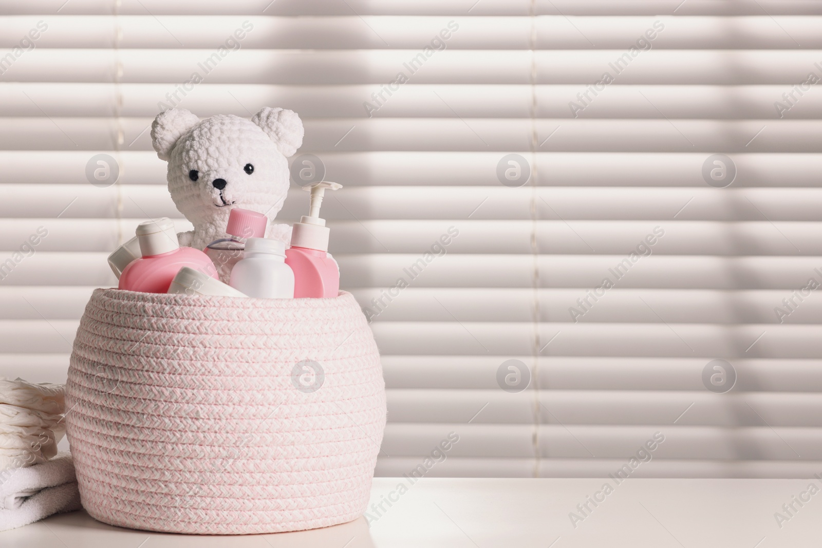 Photo of Wicker basket with baby cosmetic products and knitted toy bear on white table. Space for text