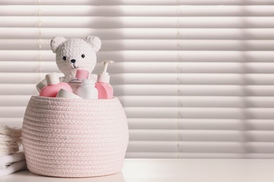 Wicker basket with baby cosmetic products and knitted toy bear on white table. Space for text