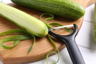 Photo of Fresh cucumbers, peels and peeler at white wooden table, closeup