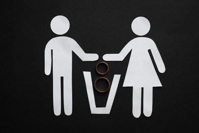 Photo of Divorce concept. Paper cutouts of couple, trash can and rings on black background, flat lay