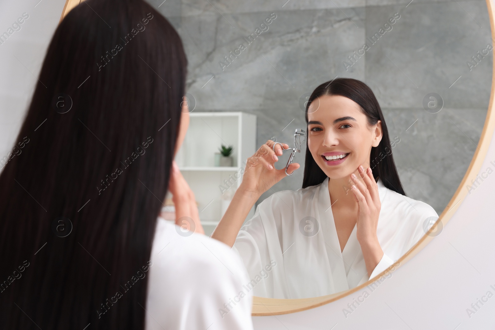 Photo of Beautiful young woman using eyelash curler near mirror in bathroom