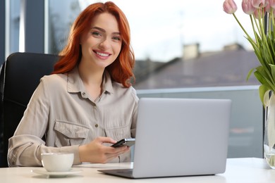 Photo of Happy woman with smartphone working on laptop at white desk in office