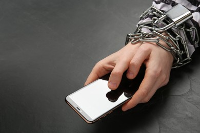 Prisoner holding smartphone in chained hands at black table, closeup. Internet addiction