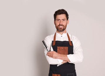 Smiling hairdresser in apron holding scissors and comb on light grey background, space for text