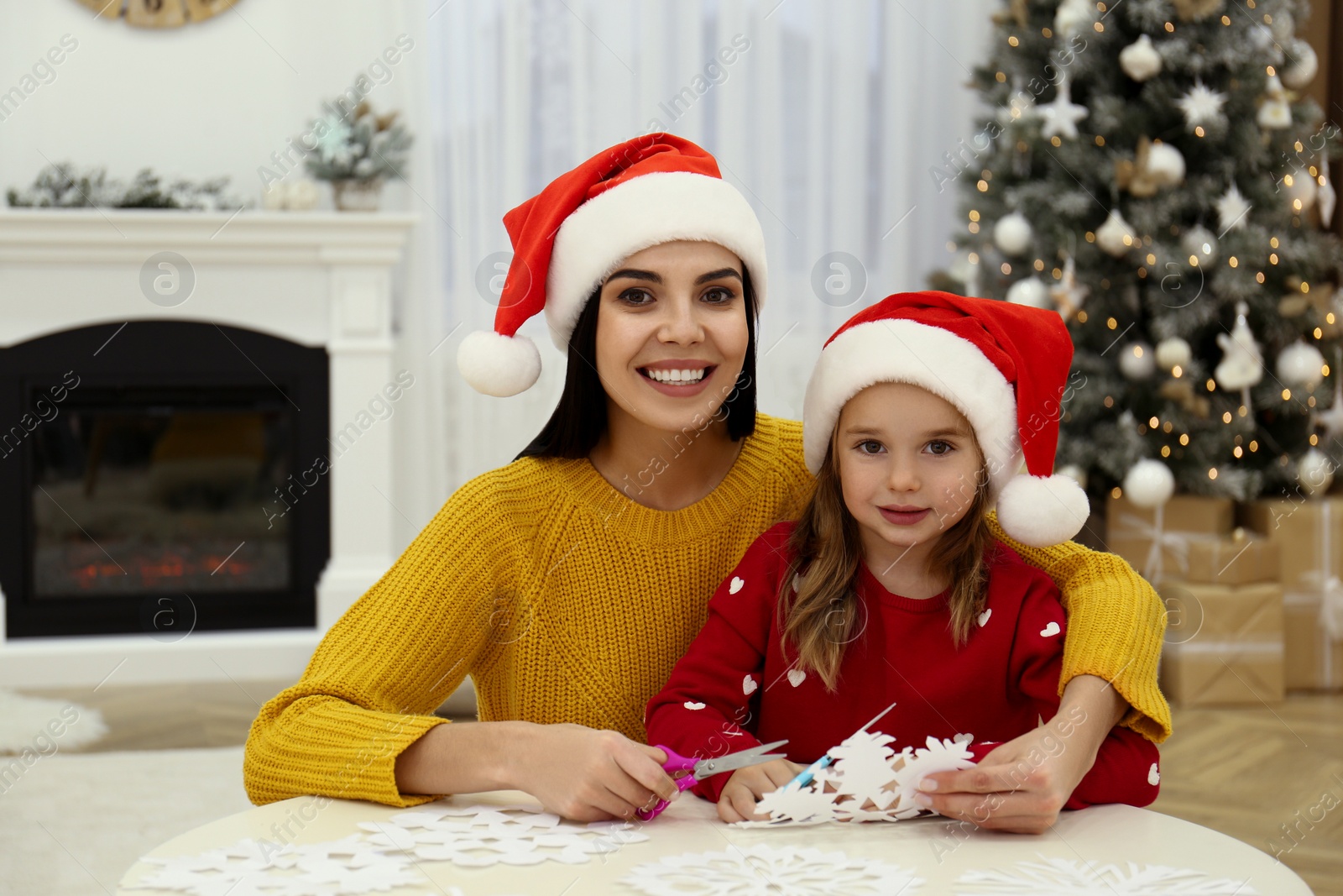 Photo of Mother and daughter in Santa hats making paper snowflake near Christmas tree at home
