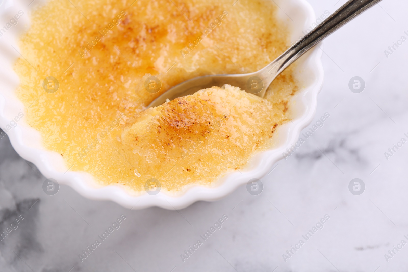 Photo of Delicious creme brulee in bowl and spoon on white marble table, top view
