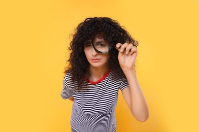 Curious young woman looking through magnifier glass on yellow background