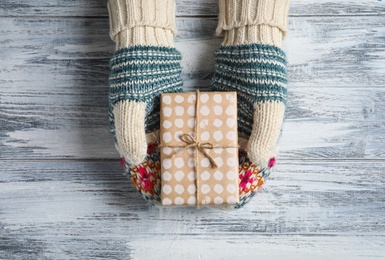 Photo of Woman wearing warm mittens holding Christmas gift on wooden background, top view