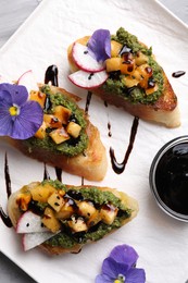 Photo of Delicious bruschettas with pesto sauce, tomatoes, balsamic vinegar and violet flowers on white plate, flat lay
