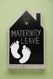 Photo of Wooden house figure with words Maternity Leave and paper cutout of baby feet on pale green background, top view