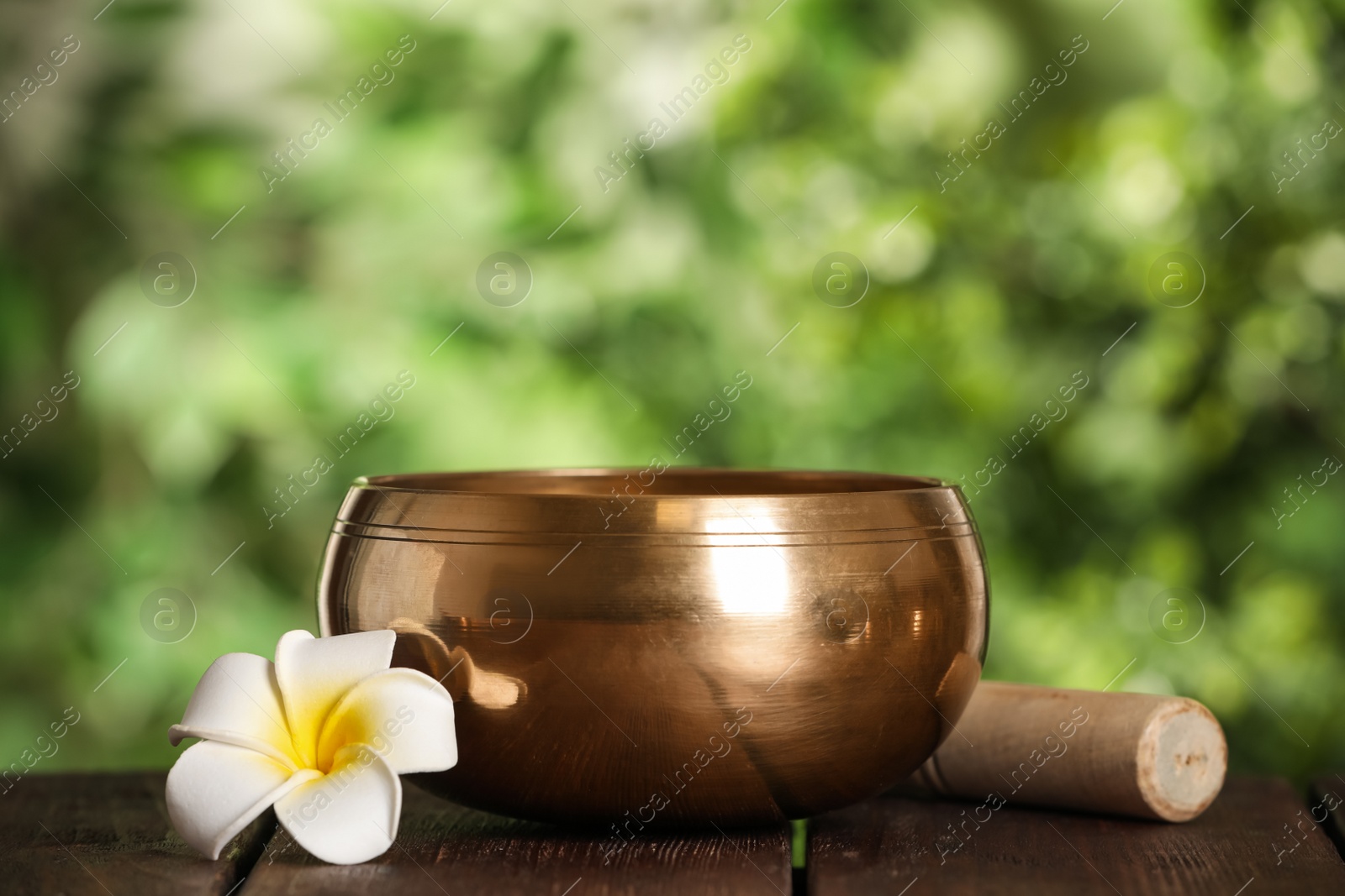 Photo of Golden singing bowl, mallet and flower on wooden table outdoors