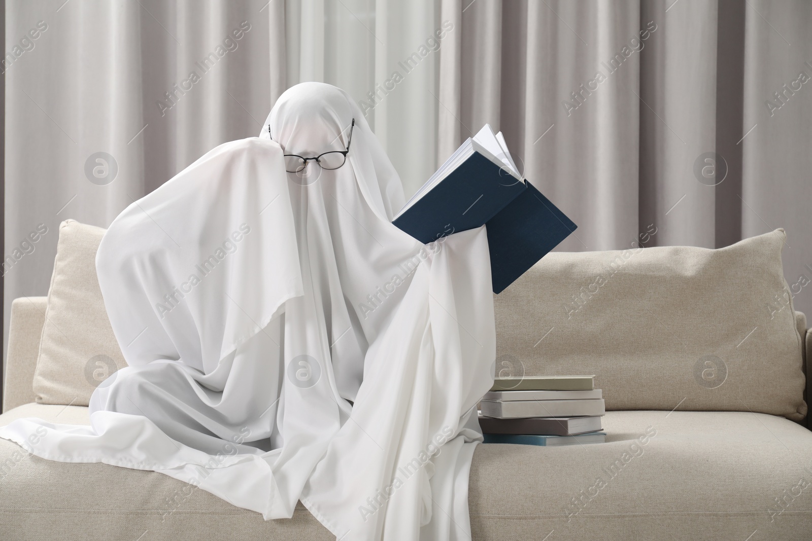 Photo of Creepy ghost. Person covered with white sheet reading book on sofa at home