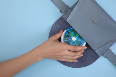 Woman putting plastic box with pills into bag on light blue background, top view