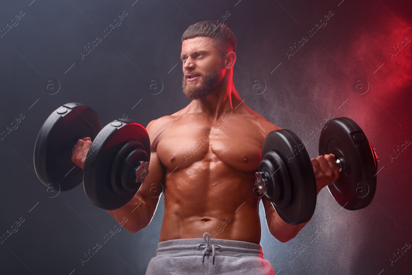 Photo of Emotional young bodybuilder exercising with dumbbells in smoke on color background, low angle view