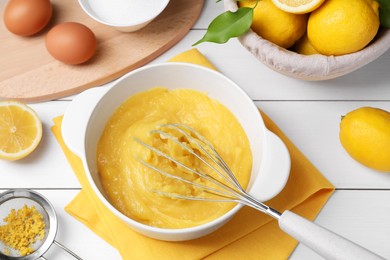 Delicious lemon curd in bowl, ingredients, whisk and sieve on white wooden table, above view