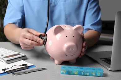 Photo of Doctor with stethoscope and piggy bank near pills at table in hospital, closeup. Medical insurance