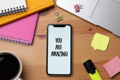 Smartphone with phrase You Are Amazing on screen and stationery against wooden background, flat lay