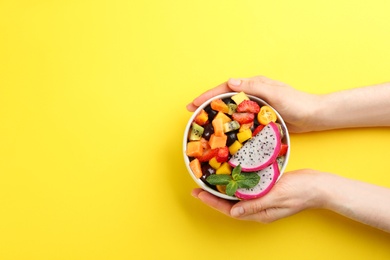 Photo of Woman with bowl of delicious exotic fruit salad on yellow background, top view. Space for text